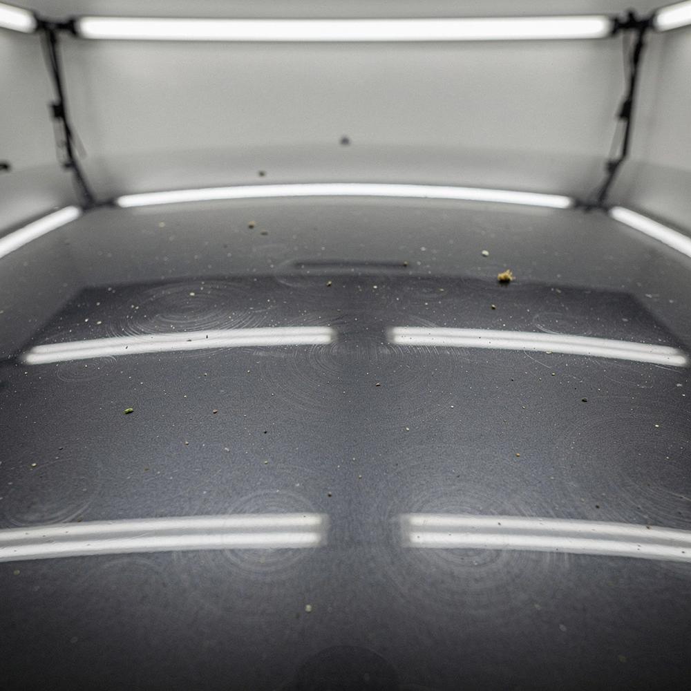 Macro view of a dark grey Cayenne hood panel before correction, showing swirl marks and dull oxidised clear coat under studio light